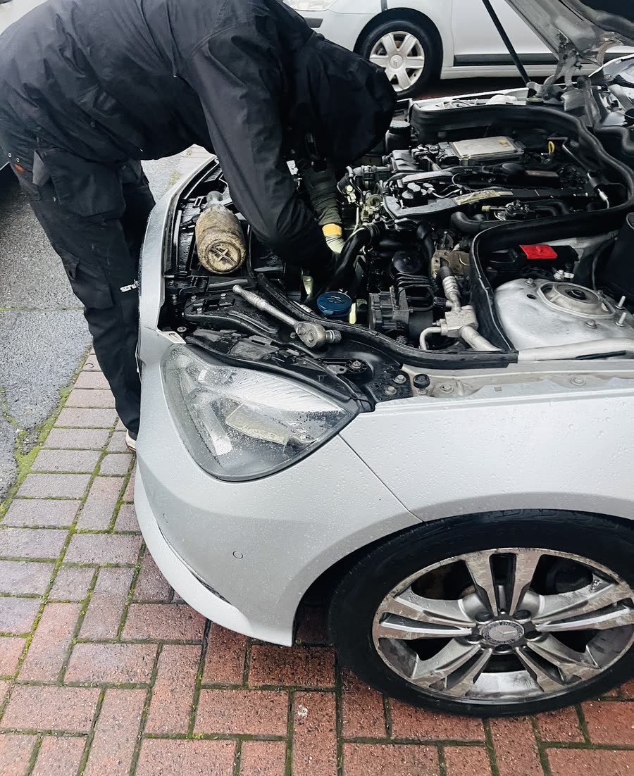 A mechanic in black workwear working under the open bonnet of a white hatchback on a block-paved driveway
