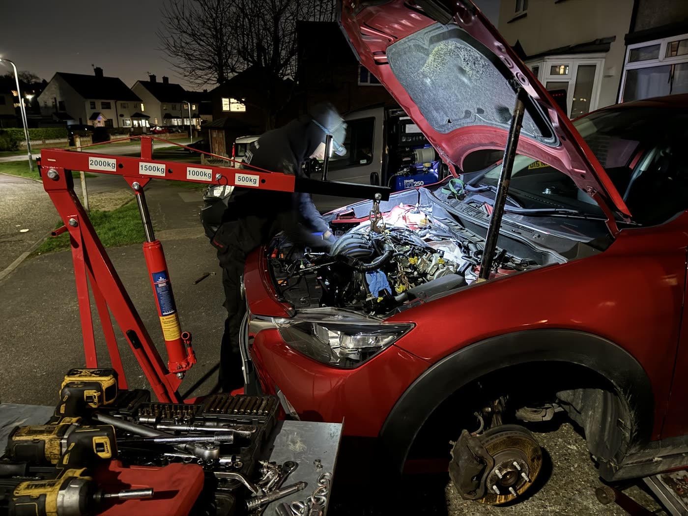 A car raised on a trolley jack at night with its bonnet up and front wheel off, tools laid out on the road beside it